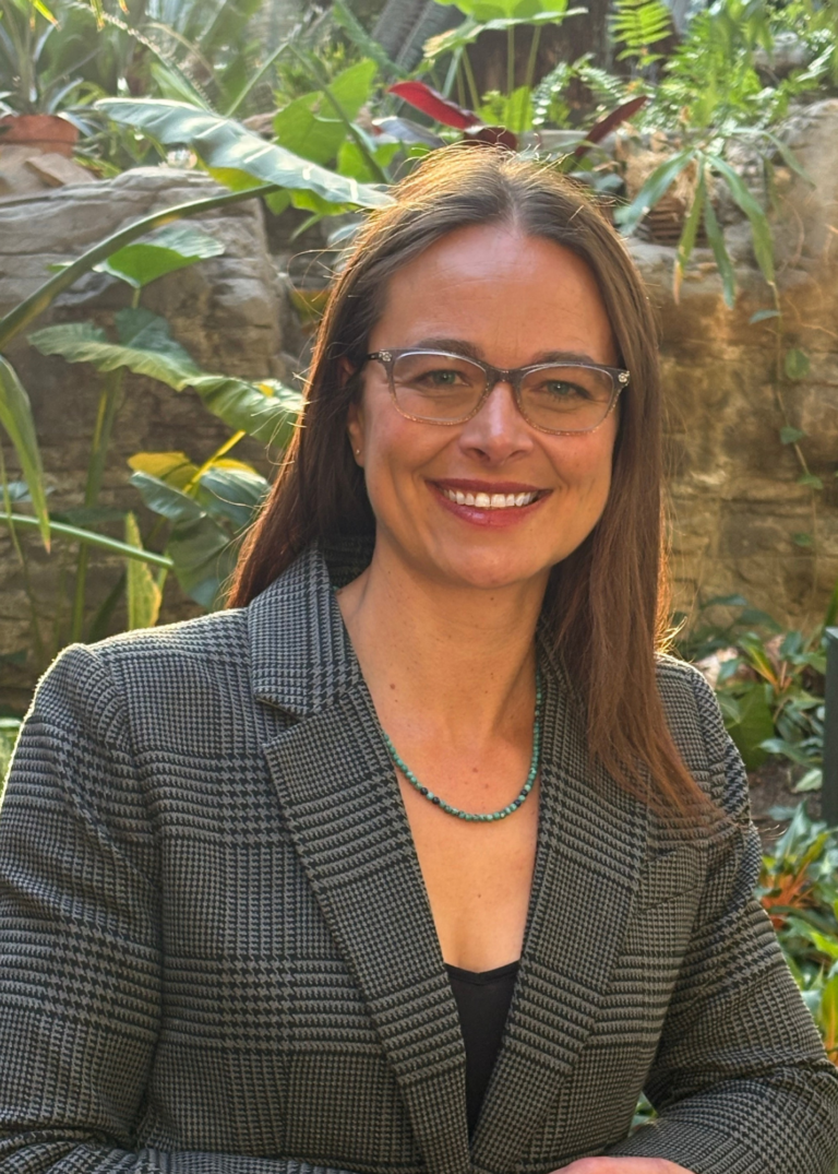 White woman, long brown straight hair, wearing grey blazer, necklace and glasses. Standing in front of green foliage.