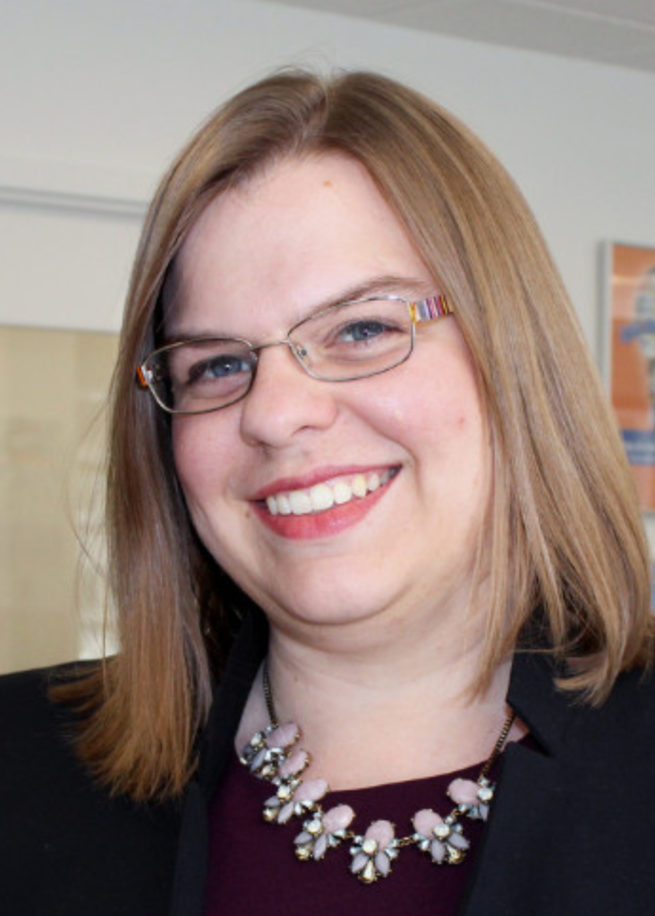 White female, dark blonde hair, clear rim glasses, in a black blazer. Chunky Necklace