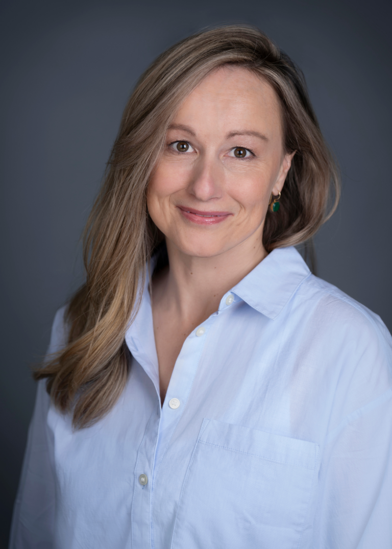 White women with a gray/blue backdrop. She has light brown hair, lipstick, and wearing a powder blue button up shirt